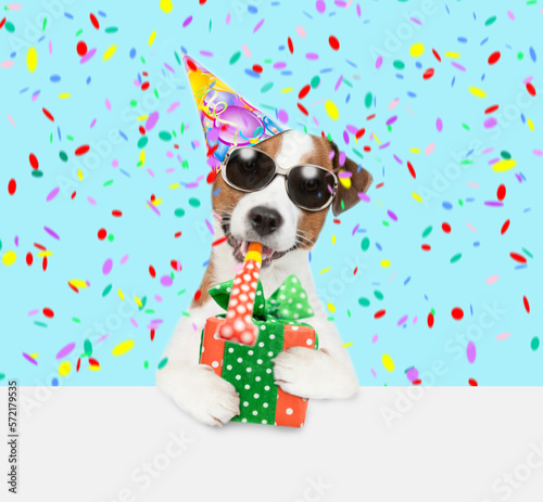 Jack russell terrier puppy wearing sunglasses and party cap blows into party horn and holds gift box above empty white banner © Ermolaev Alexandr