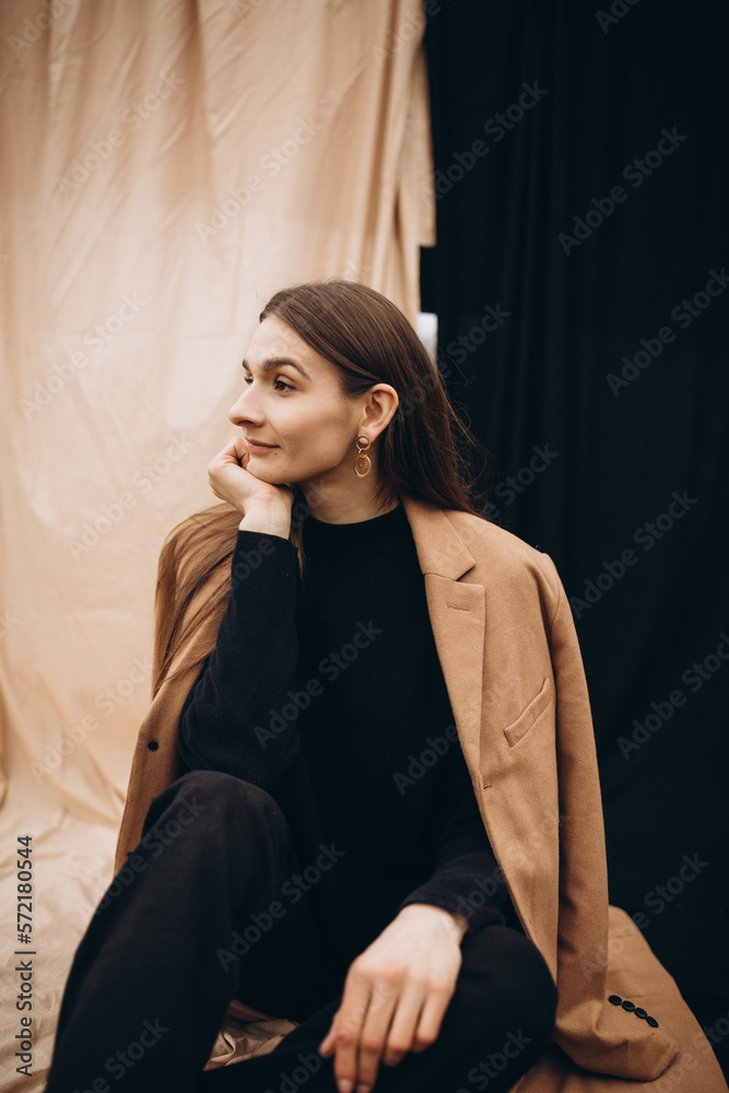 beautiful girl in a black sweater and black pants and a beige coat on a black and beige fabric background. cold cloudy weather and dry yellow tall grass and a girl. stylish fashion portrait. beautiful girl in a black sweater and black pants and a beige coat on a black and beige fabric background. cold cloudy weather and dry yellow tall grass and a girl. stylish fashion portrait.