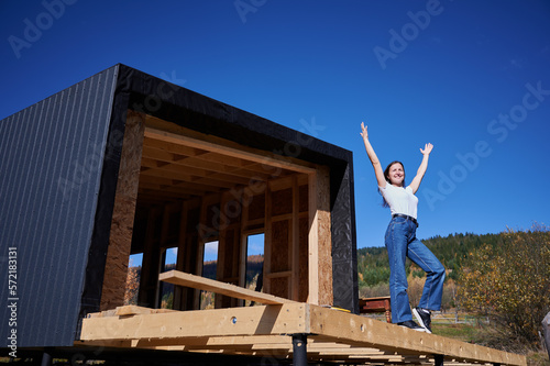 Happy woman on construction site standing on terrace at incomplete wooden frame house on sunny day with hands up.