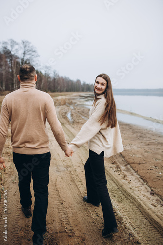 a man and a woman are walking along the shore of the lake, cold foggy weather. romantic photo in beige tones. holding a woman's hand, a love story. beige sweaters. romantic photos by the lake © Anhelina Tyshkovets