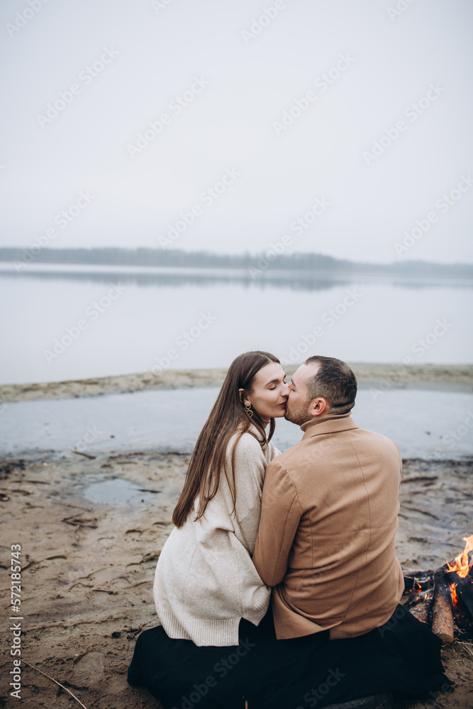 foggy cold evening couple in love sitting by the fire and kissing. a campfire on the shore of a lake and a man and a woman sitting. clothes in beige tones. a love story in cold foggy weather
