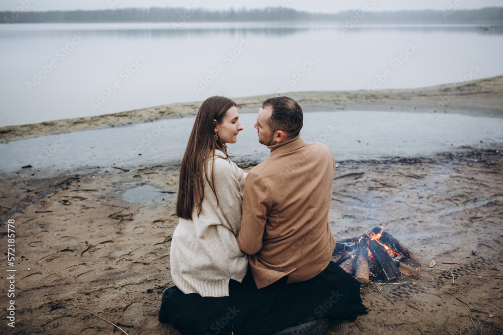 foggy evening by the lake, a bonfire is burning. a man and a woman are sitting on the shore of a lake near a bonfire. beige men's coat. love story photo shoot in cloudy foggy weather