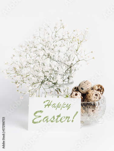 Fototapeta Naklejka Na Ścianę i Meble -  White gypsophila in vase, words Happy Easter on white sheet, quail eggs in crystal bowl on white.