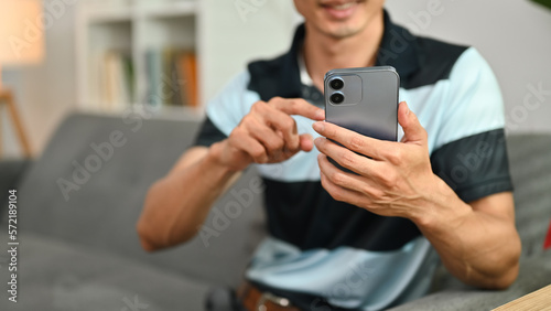 Cropped shot of smiling asian man sitting on couch reading messages on mobile phone. Selected focus on hands
