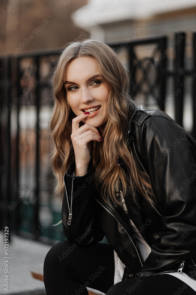 Fototapeta premium Close up portrait of a beautiful smiling young blonde woman in a black leather jacket on the a city street background