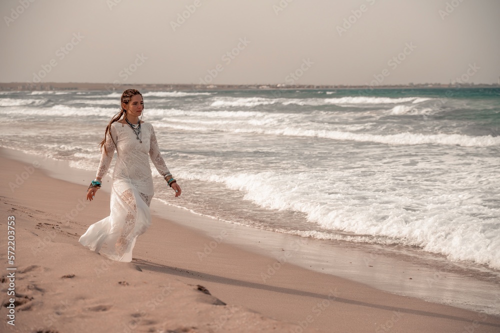 Model in boho style in a white long dress and silver jewelry on the beach. Her hair is braided, and there are many bracelets on her arms. Model in boho style in a white long dress and silver jewelry on the beach. Her hair is braided, and there are many bracelets on her arms.