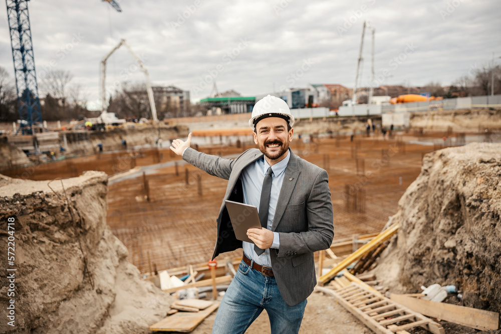 A proud engineer is standing on construction site with tablet in his hands and showing at the site with foundation.