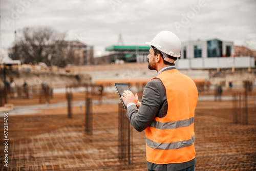 Back view of a site manager standing on site and scrolling on the tablet while supervising works.