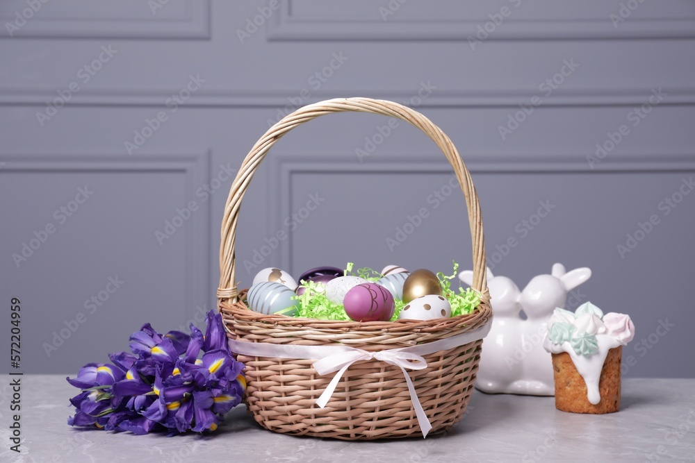 Easter basket with many painted eggs near tasty cake, flowers and figure of rabbits on grey textured table