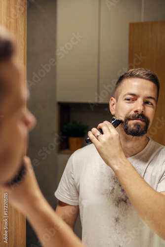 Portrait of a bearded man styling his beard with shaver in bathroom.