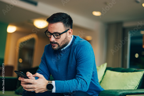 Focused businessman with glasses typing on the phone, wearing a blue sweater, and sitting on the couch.