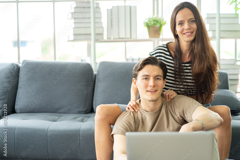 A couple sitting together in the living room enjoy relaxing at home. Young man sits on floor while his girlfriend sits on couch watching online movie from laptop computer. Couple leisure activity A couple sitting together in the living room enjoy relaxing at home. Young man sits on floor while his girlfriend sits on couch watching online movie from laptop computer. Couple leisure activity