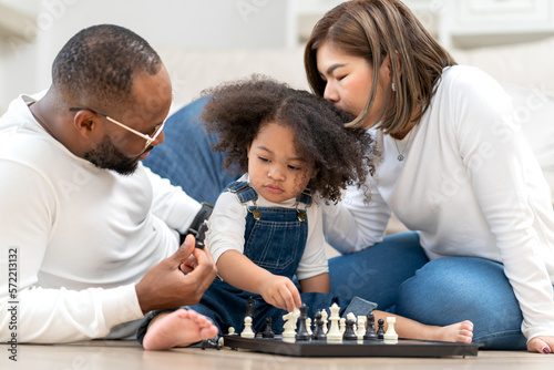 Cute little girl playing chess board as her father sitting by. Multiracial family happy leisure together with daughter kid at home. Multiethnic child has fun playing chess pieces with her dad and mom.