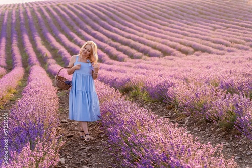 Fototapeta Naklejka Na Ścianę i Meble -  Woman lavender field sunset. Romantic woman walks through the lavender fields. illuminated by sunset sunlight. She is wearing a blue dress with a hat.