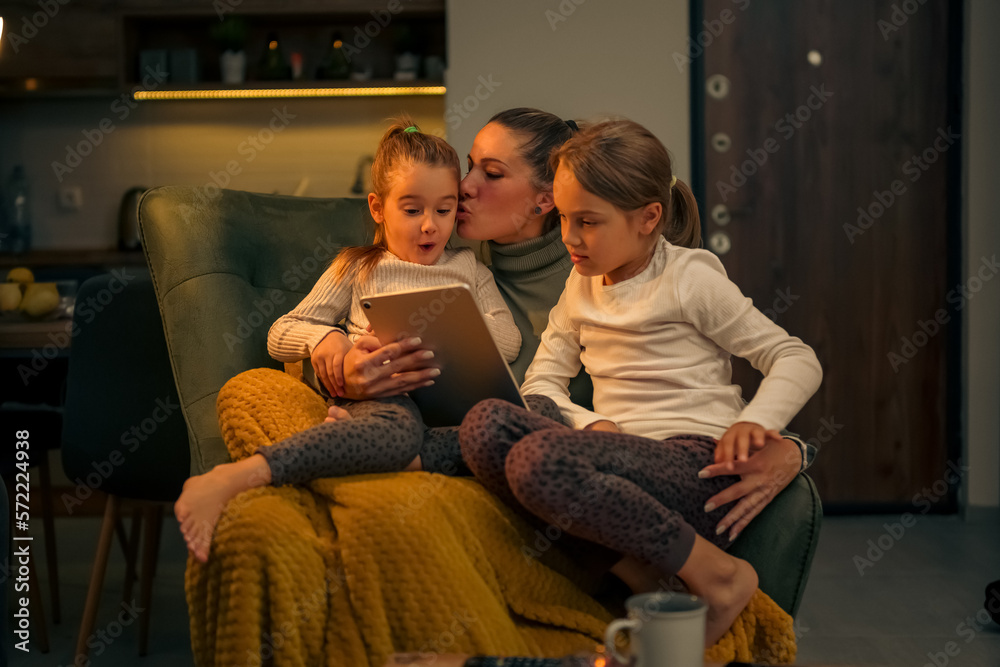 Mother is kissing her daughter, her and her daughters are comfortable on an armchair watching videos on a tablet, they are happy