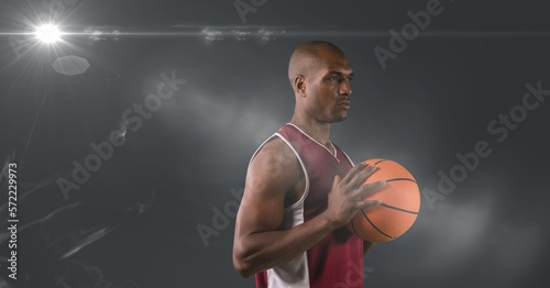 African american male basketball player holding basketball against light spot on grey background