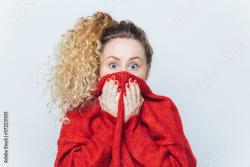 Shocked scared beautiful female stares at camera, being afraid of something awful, covers face with collar, dressed in red loose sweater, isolated over white background. Fear and fright concept photo