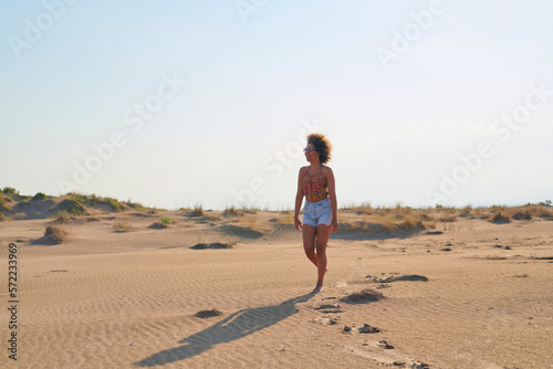 Full length portrait of happy beautiful woman with afro hairstyle in sunglasses, top and denim shorts walking in sunny desert on summer day. She is looking away with excitement.