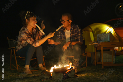 Asian couple roasting marshmallows together at a campfire where they set up a tent to camp by the lake at night.