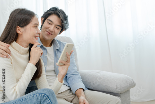 Asian couple smiling and happily use smartphone to online shopping. Husband ready to pay birthday present for beautiful wife. Find accommodation online for honeymoon, App online, book flight tickets.