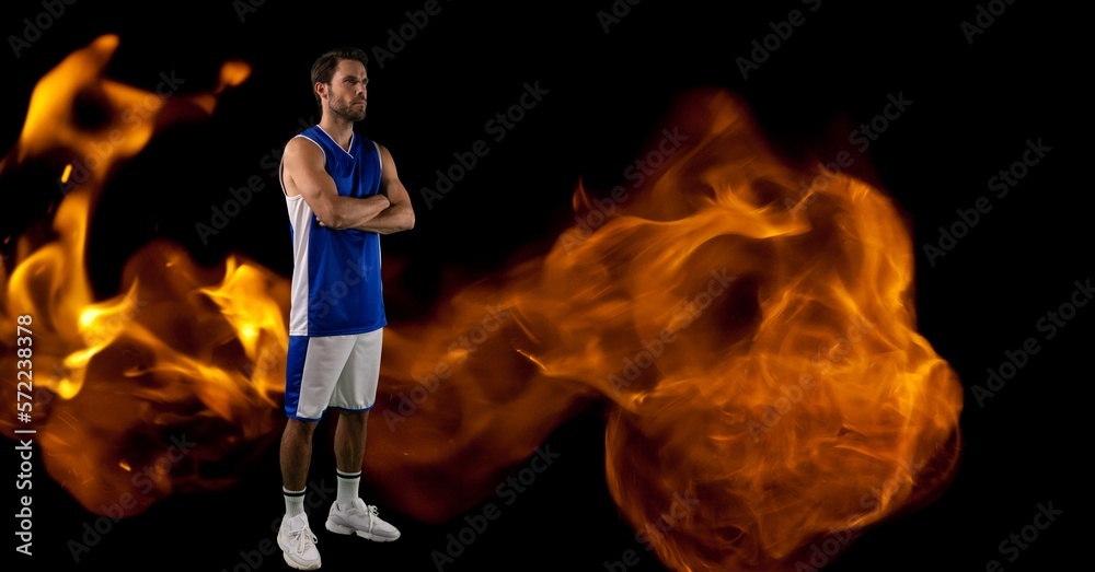 Composition of male basketball player standing with arms crossed over flames on black background