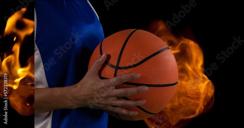 Composition of midsection of male basketball player holding ball over flames on black background