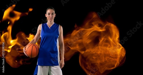 Composition of female basketball player standing holding ball over flames on black background