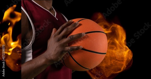 Composition of midsection of male basketball player holding ball over flames on black background