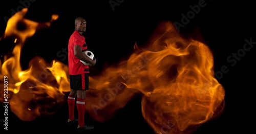 Composition of male football player standing holding ball over flames on black background