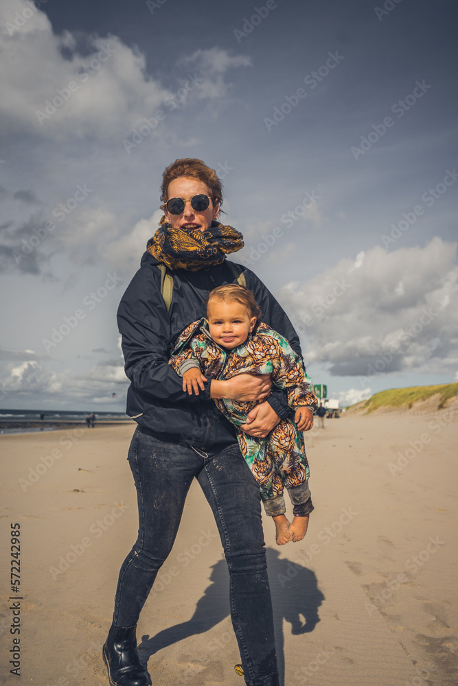 Mutter mit Kleinkind auf dem Arm ab einem Strand an der Ostsee, Wetter ist frisch und der Wind geht stark, trotzdem sind beide fröhlich und lachen 