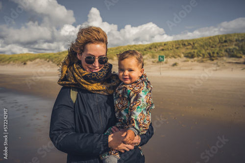 Mutter mit Kleinkind auf dem Arm ab einem Strand an der Ostsee, Wetter ist frisch und der Wind geht stark, trotzdem sind beide fröhlich und lachen 