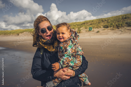 Mutter mit Kleinkind auf dem Arm ab einem Strand an der Ostsee, Wetter ist frisch und der Wind geht stark, trotzdem sind beide fröhlich und lachen 