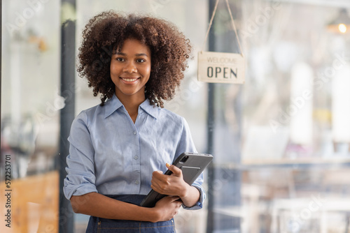 Successful african woman in apron standing coffee shop door. Happy small business owner holding tablet and working. Smiling portrait of SME entrepreneur seller business standing with copy space.