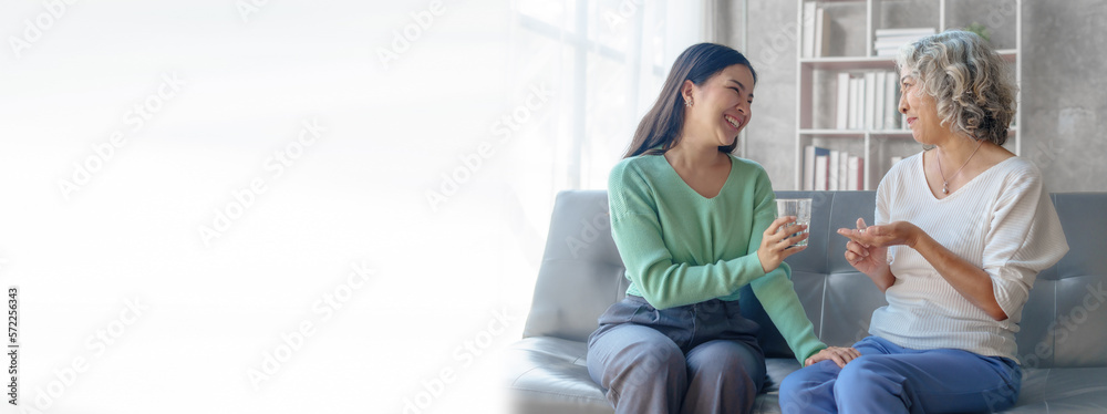 Obraz premium 60s asian mother elderly sitting on sofa with young asia female daughter together in living room. give medicine and water
