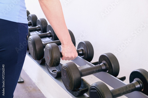 Woman in the gym takes a dumbbell from the rack. Black metal dumbbells of different weights on a rack in a gym. Sports equipment for increasing muscle mass. The concept of doing sports.