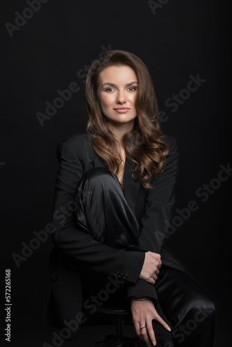 Fashion and make-up concept. Portrait of beautiful brunette woman with black suit and big wavy hair in dark studio background. Model sitting on chair and hugging her one leg. Woman looking at camera