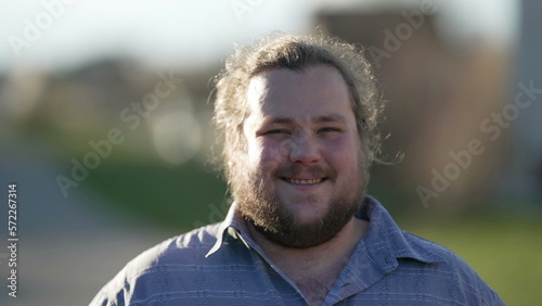 One happy overweight young man smiling at camera standing outdoors in sunlight. Natural casual male caucasian person looking at camera