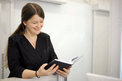 A young woman sits at a computer at the reception of a white light office hospital dentistry long hair black clothes white computer laptop hands of black notebook with notes notes interesting book