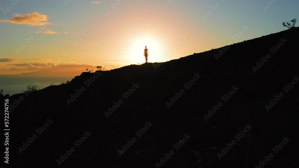 Silhouette of a person on side of mountain at orange sunset sky. Hiker woman standing on highland with ocean view. Sunlight illuminates dark nature. Silhouette of a person on side of mountain at orange sunset sky. Hiker woman standing on highland with ocean view. Sunlight illuminates dark nature.