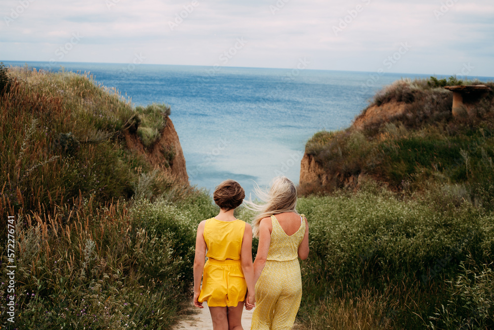 Mom and daughter in yellow summer clothes walk hand in hand through the green hills to the seashore, in the afternoon on a sunny day with wind.