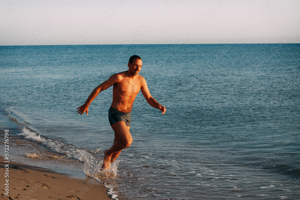 A slender muscular tanned guy in blue swimming trunks runs along the sandy shore of the ocean and smiles on a bright sunny day.