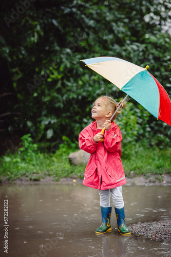 Fototapeta Naklejka Na Ścianę i Meble -  A little beautiful girl blonde, in boots and with umbrella walking along the puddles in the rain. Happy child. A walk under the summers of the rain.