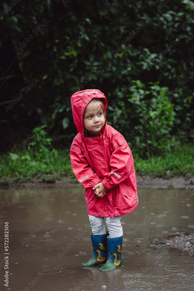 Beautiful girl walking along the puddles under the summer rain in a water-repellent raincoat and boots, on a green background of trees and grass. Carefree childhood. Happy child. Beautiful girl walking along the puddles under the summer rain in a water-repellent raincoat and boots, on a green background of trees and grass. Carefree childhood. Happy child.