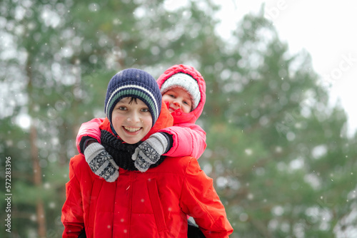 Portrait of two children, smiling teenage boy standing outside in forest in snowy winter, holding little girl on back.