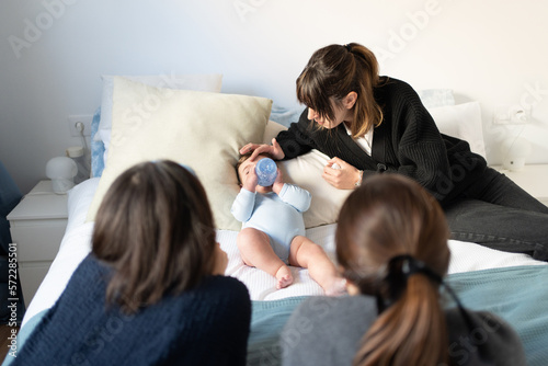 baby taking bottle with mother caressing, mother's friends looking on with loving face, new birth on girl in her twenties. First little one among the group of friends