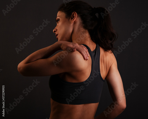 Foto Female sporty muscular with ponytail doing stretching workout of the shoulders, blades in sport bra, holing the neck the hand on dark grey background with empty copy space