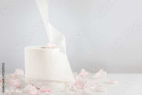 Roll of natural white toilet paper on a light background with rose petals. White toilet tissue, Hygiene product. Restroom soft touch toilet paper. Soft focus style image. Front view, copy space