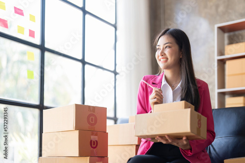Young woman, an Asian online business owner, smiles, prepares parcel boxes and checks online orders for products to be delivered to customers © Natee Meepian