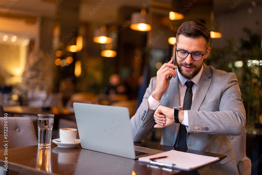 Handsome confident businessman in suit talking on smartphone, looking at hand watch, checking time, sitting in modern restaurant waiting for business partner to come on business dinner. Handsome confident businessman in suit talking on smartphone, looking at hand watch, checking time, sitting in modern restaurant waiting for business partner to come on business dinner.