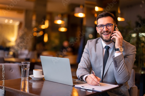 Smiling modern businessman talking to business partner on smartphone, writing down ideas or future business plan, taking notes on clipboard while working and having coffee in restaurant in the evening